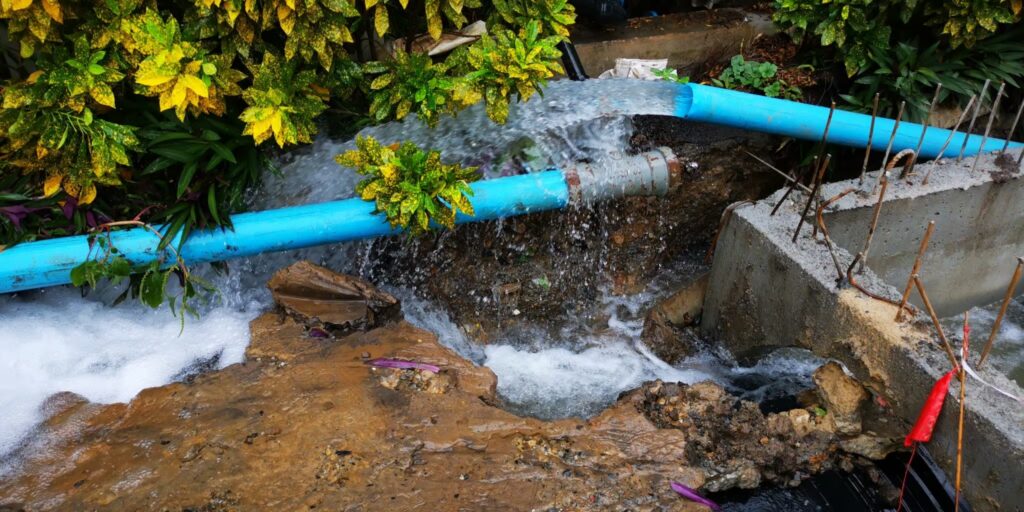 Pipe repair in Dallas, TX, showing a blue pipe with water leaking amidst greenery and rocky terrain, illustrating plumbing maintenance and environmental considerations.